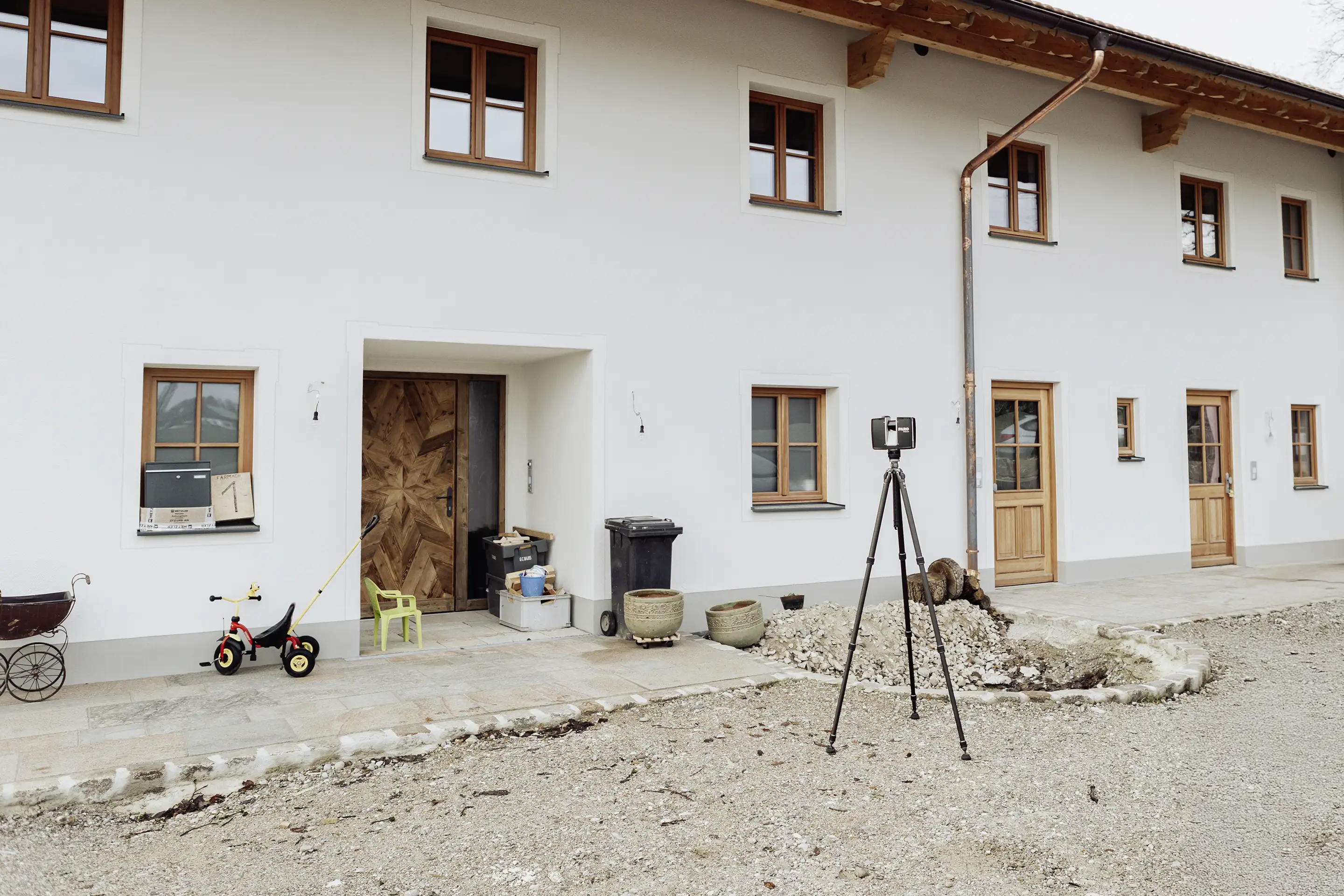 Ein 3D-Laserscanner steht auf einem Stativ vor einem Wohnhaus mit Holzfenstern und heller Fassade, um die Gebäudeaußenmaße präzise zu erfassen.