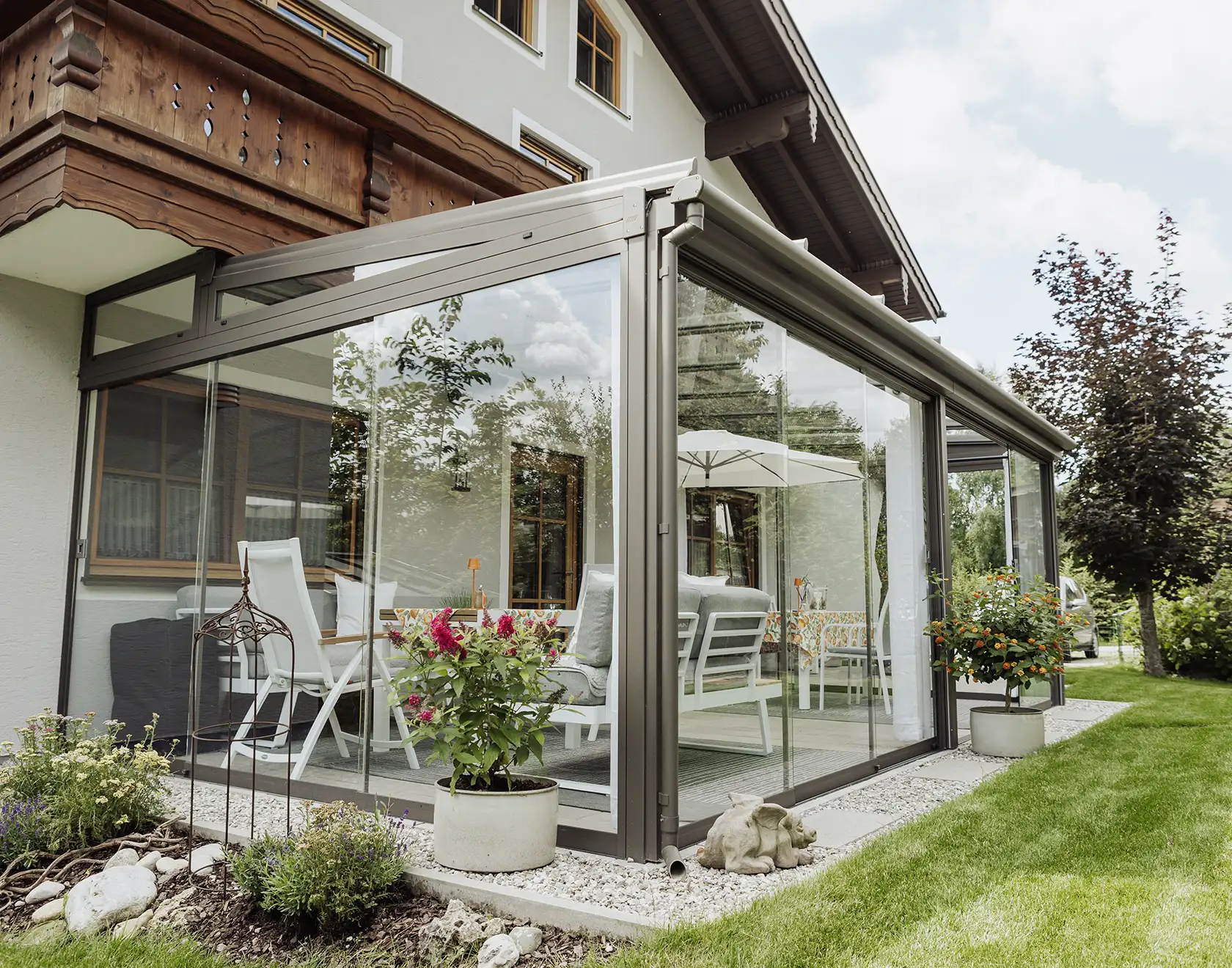 Ein moderner Wintergarten aus Glas und Metall ist an ein Wohnhaus mit Holzverkleidung angebaut und dient als Erweiterung des Wohnraums.
