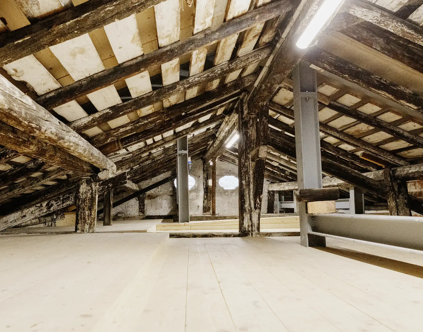Ein alter Dachboden mit sichtbaren Holzbalken wird saniert; neue Holzböden und Metallstützen stabilisieren die Struktur.