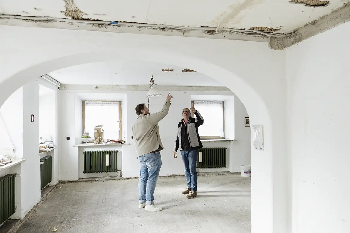 Zwei Personen begutachten gemeinsam die Decke eines renovierungsbedürftigen Raumes, um den Sanierungsbedarf zu ermitteln.