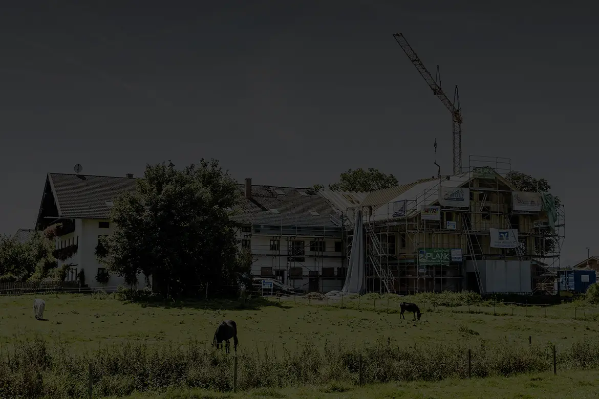 Ein landwirtschaftliches Gebäude im Bau mit Baukran und Holzrahmenkonstruktion; im Vordergrund weiden Kühe auf der Wiese.