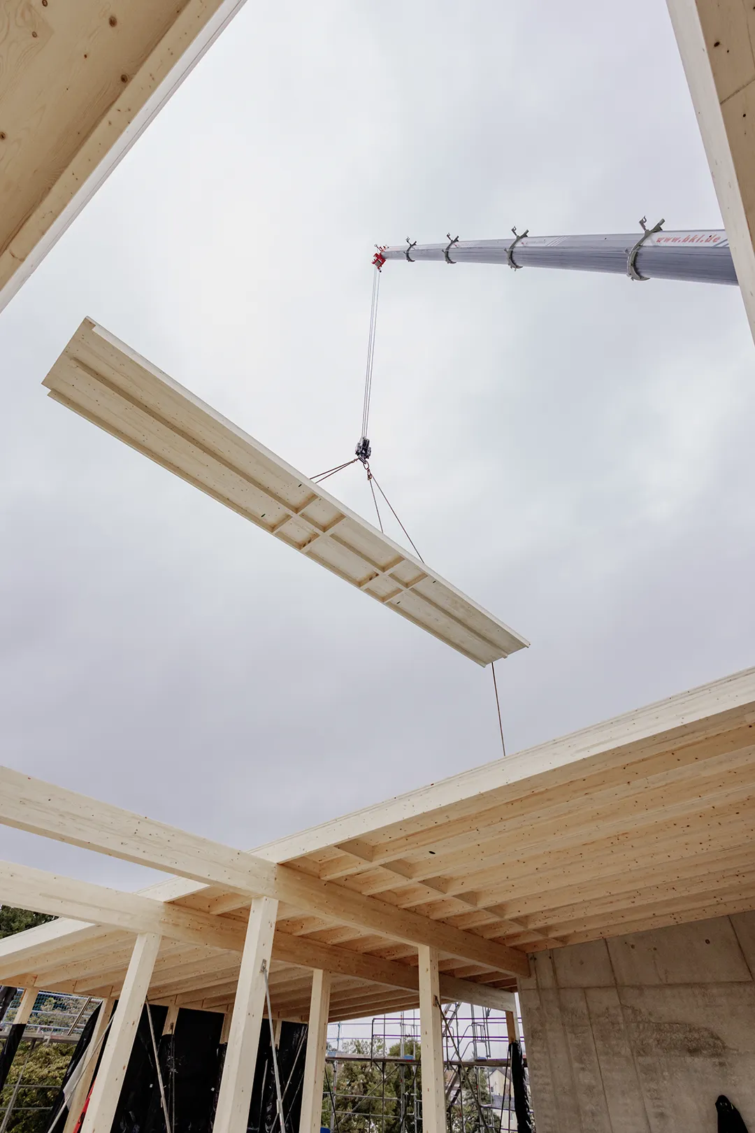 Ein Kran hebt einen großen Holzbalken auf eine Holzrahmenbaustelle bei bedecktem Himmel.