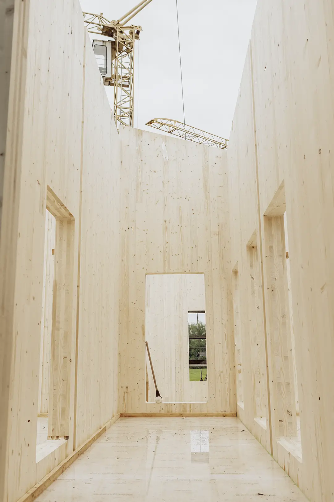 Innenansicht eines im Bau befindlichen, noch unfertigen Holzgebäudes mit hohen, holzgetäfelten Wänden und einem darüber sichtbaren Baukran.