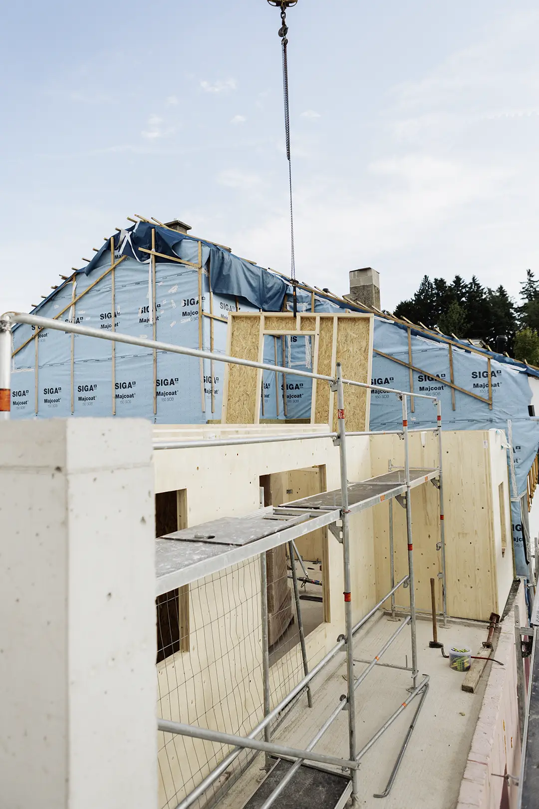 Neben einem im Bau befindlichen Holzhaus ist ein Gerüst aufgebaut, dessen Dachstuhl mit blauer Wetterschutzfolie umwickelt ist, während ein Kran eine Wandplatte anhebt.