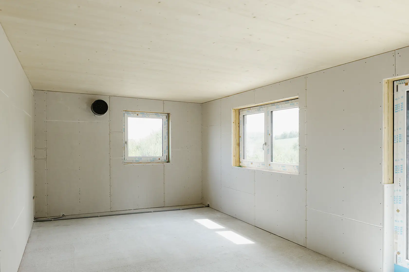 Unfertiger Raum mit Gipskartonwänden, zwei mit Plastikfolie abgedeckten Fenstern, einer CLT-Decke und einem Betonboden.