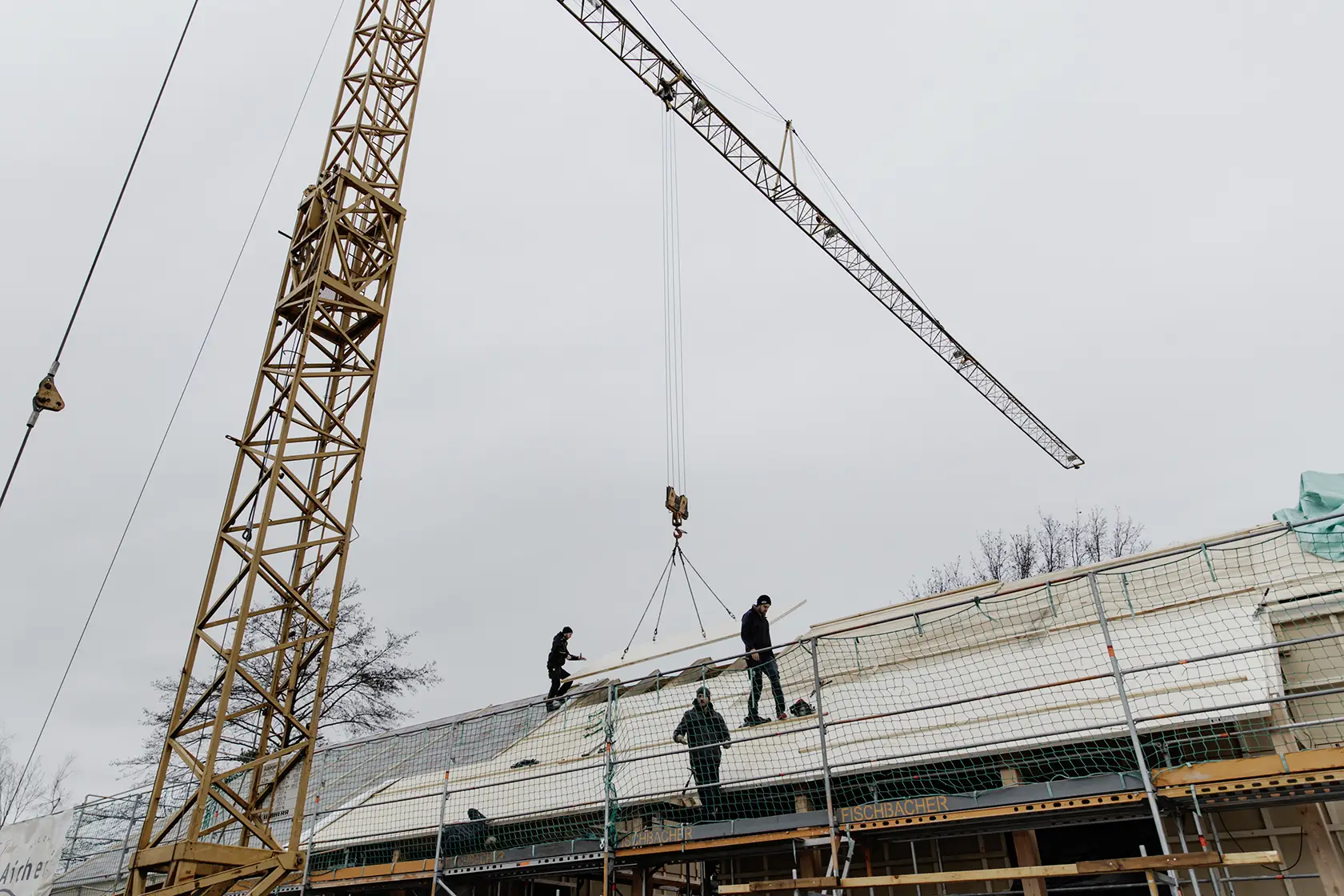 Bauarbeiter helfen auf einem schrägen Dach beim Anheben eines großen CLT-Dachs durch einen gelben Kran an einem bewölkten Tag.