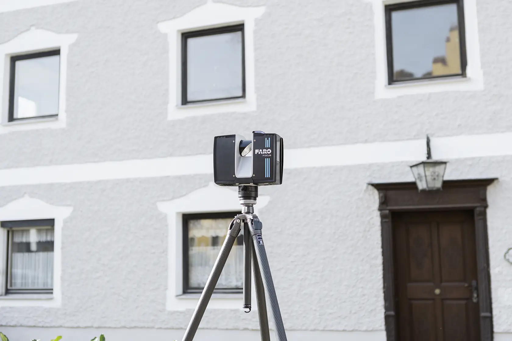 Ein FARO Focus 3D-Laserscanner ist auf einem Stativ vor einem Gebäude mit weiß gerahmten Fenstern und einer braunen Tür montiert.