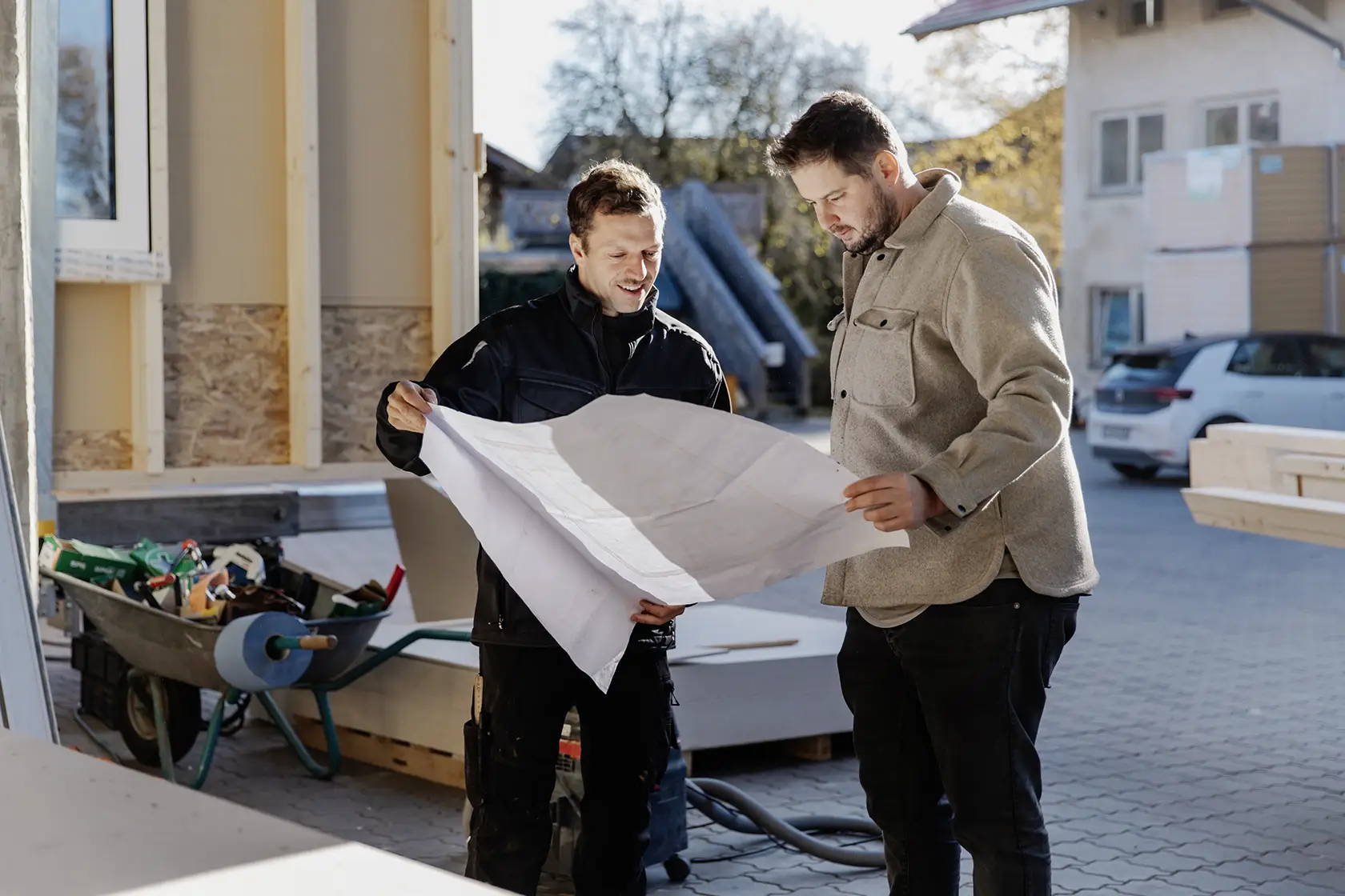 Zwei Männer prüfen auf einer Baustelle Baupläne; im Hintergrund sind Werkzeuge und Baumaterialien zu sehen.