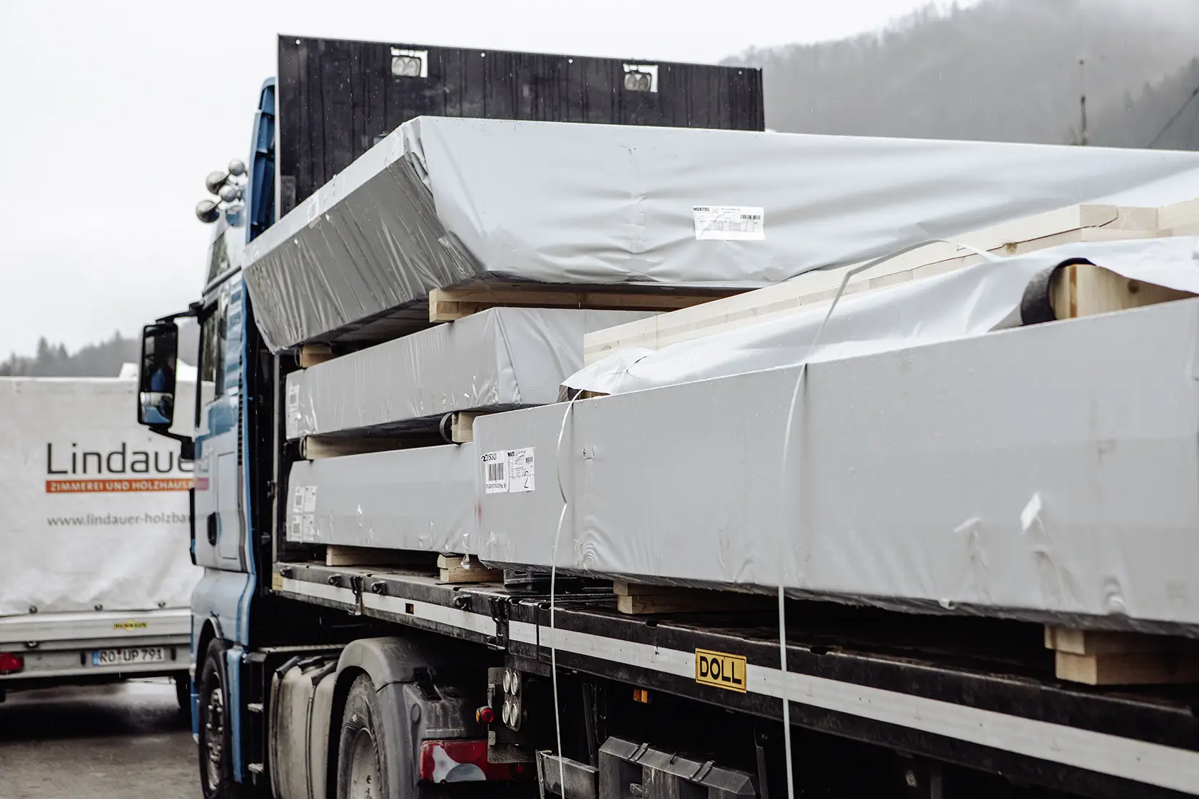 Ein mit großen, verpackten Baumaterialien beladener Pritschenwagen parkte neben einem anderen Fahrzeug mit einem Lindauer-Firmenschild.