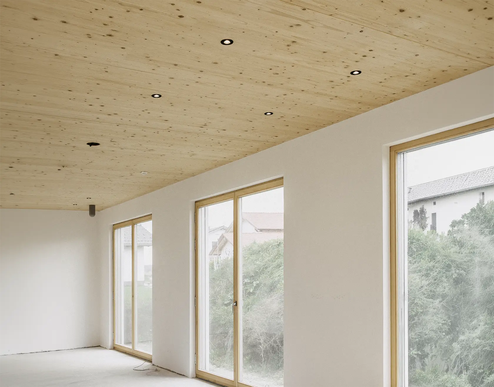 Leerer, geräumiger Raum mit großen, bodentiefen Fenstern in hellem Holzrahmen und einer Holzdecke mit Einbauleuchten.
