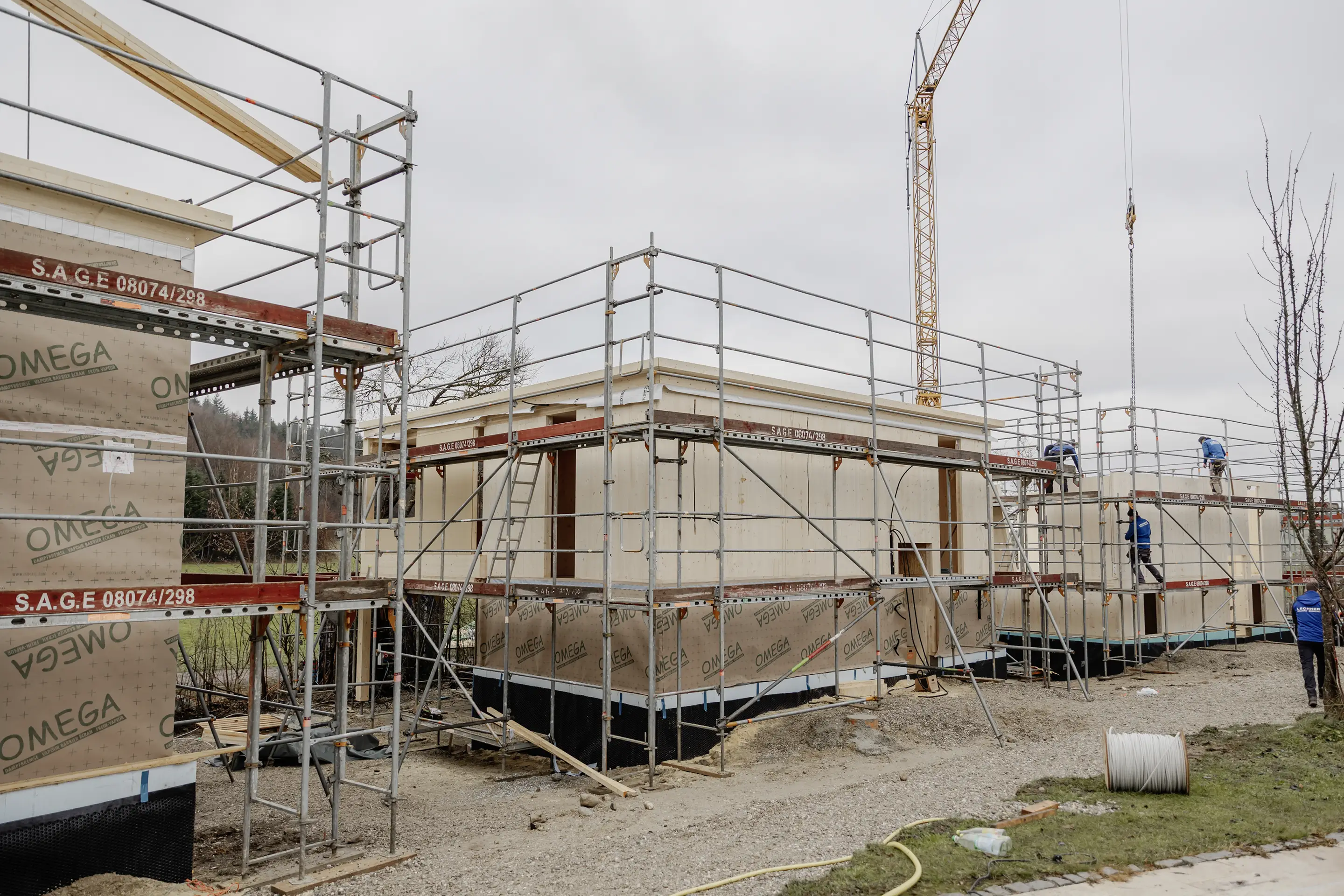 Eine Baustelle, auf der Arbeiter unter bewölktem Himmel hölzerne Gebäudegerüste montieren, umgeben von einem Metallgerüst und einem hohen Kran.