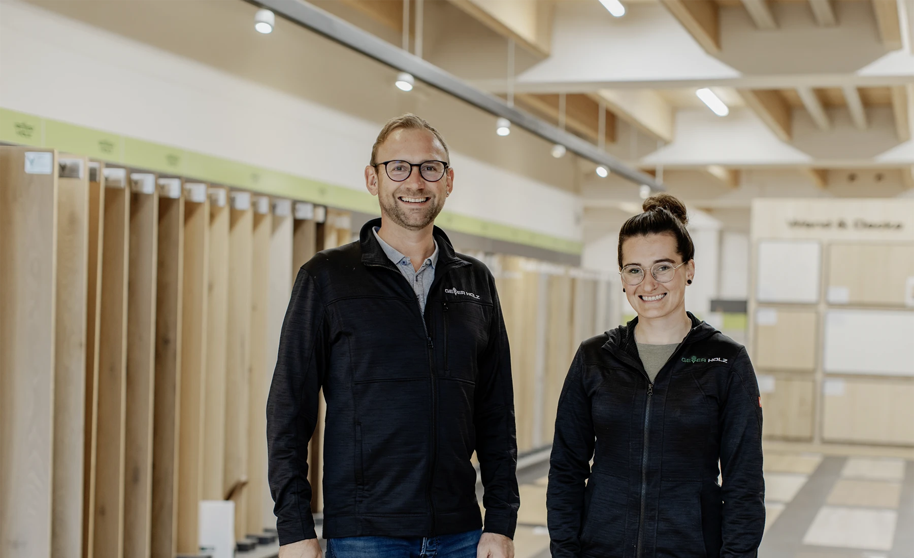 Ein lächelnder Mann und eine lächelnde Frau in schwarzen Jacken stehen in einem Ausstellungsraum für Holzfußböden.