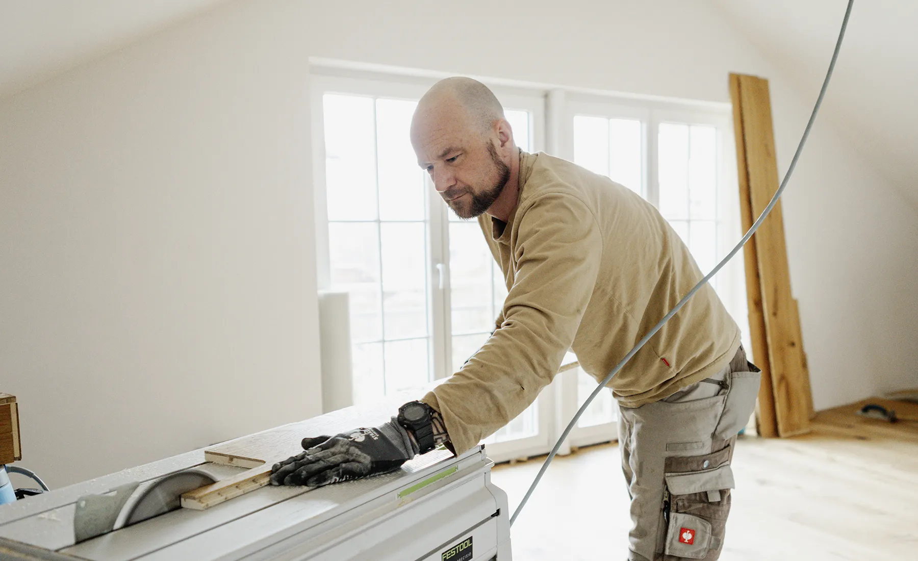 Ein Mann mit Handschuhen bedient eine Festool-Tischkreissäge in einem hellen Raum; im Hintergrund sind Holzplanken zu sehen.