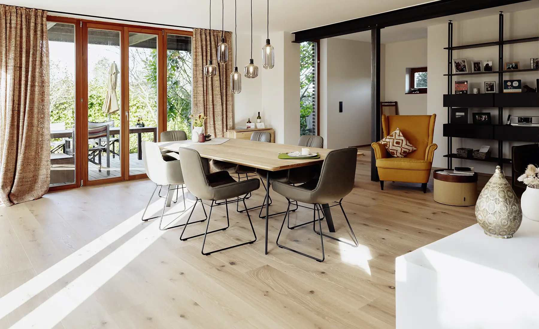 Heller Essbereich mit einem Holztisch, umgeben von sechs modernen Stühlen, Pendelleuchten, einem senffarbenen Sessel und großen Glastüren, die sich zu einer Außenterrasse öffnen.