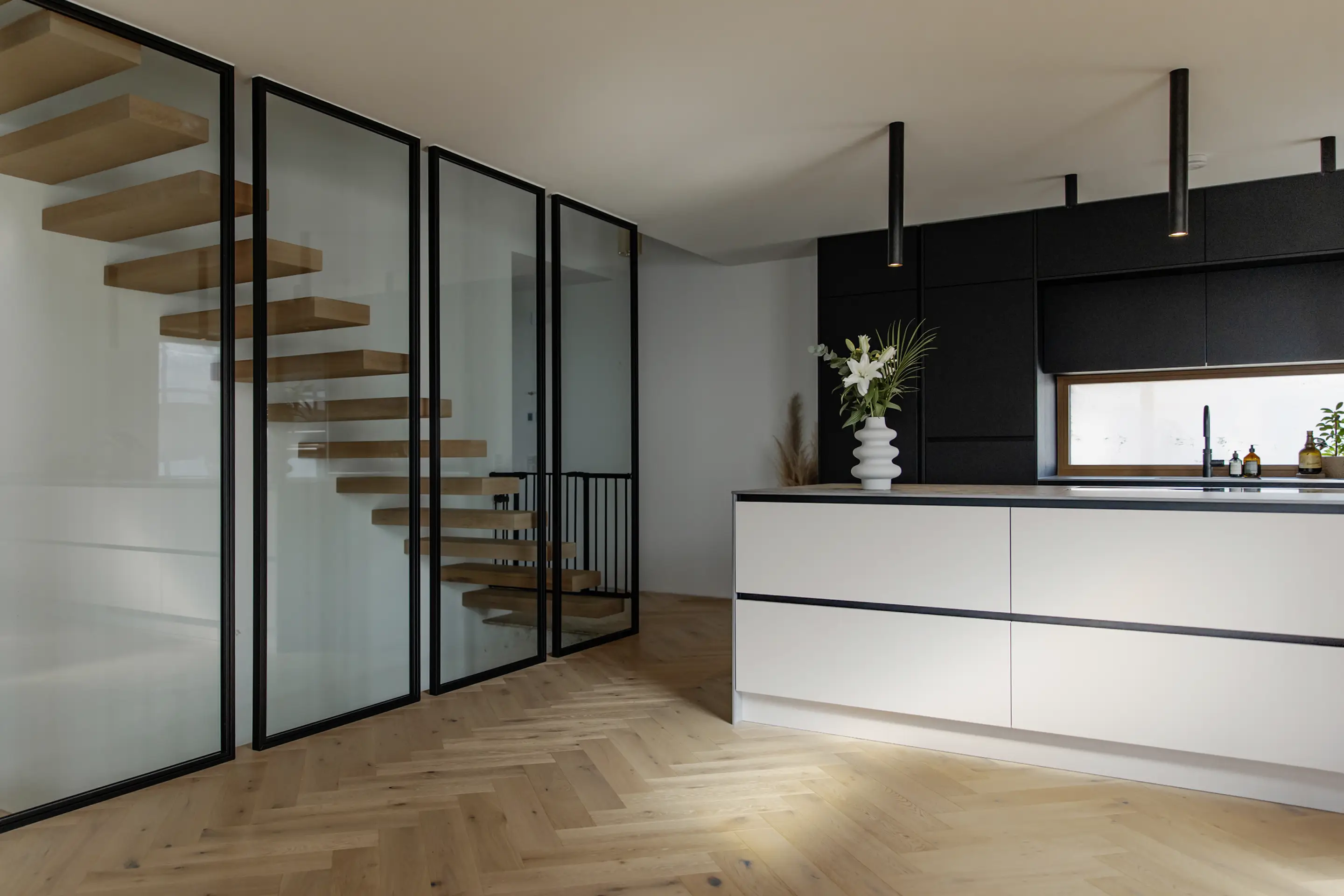 Moderne Küche mit weißer Kochinsel, schwarzen Schränken, freitragender Holztreppe hinter Glaspaneelen und einer Vase mit weißen Blumen auf der Kochinsel.