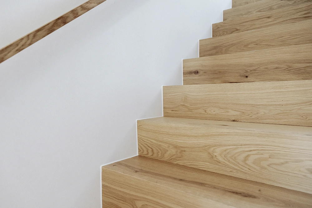 
Close-up view of light wood stairs with natural grain and a simple wooden handrail beside a white wall.
Nahaufnahme einer hellen Holztreppe mit natürlicher Maserung und einem schlichten Holzhandlauf neben einer weißen Wand.