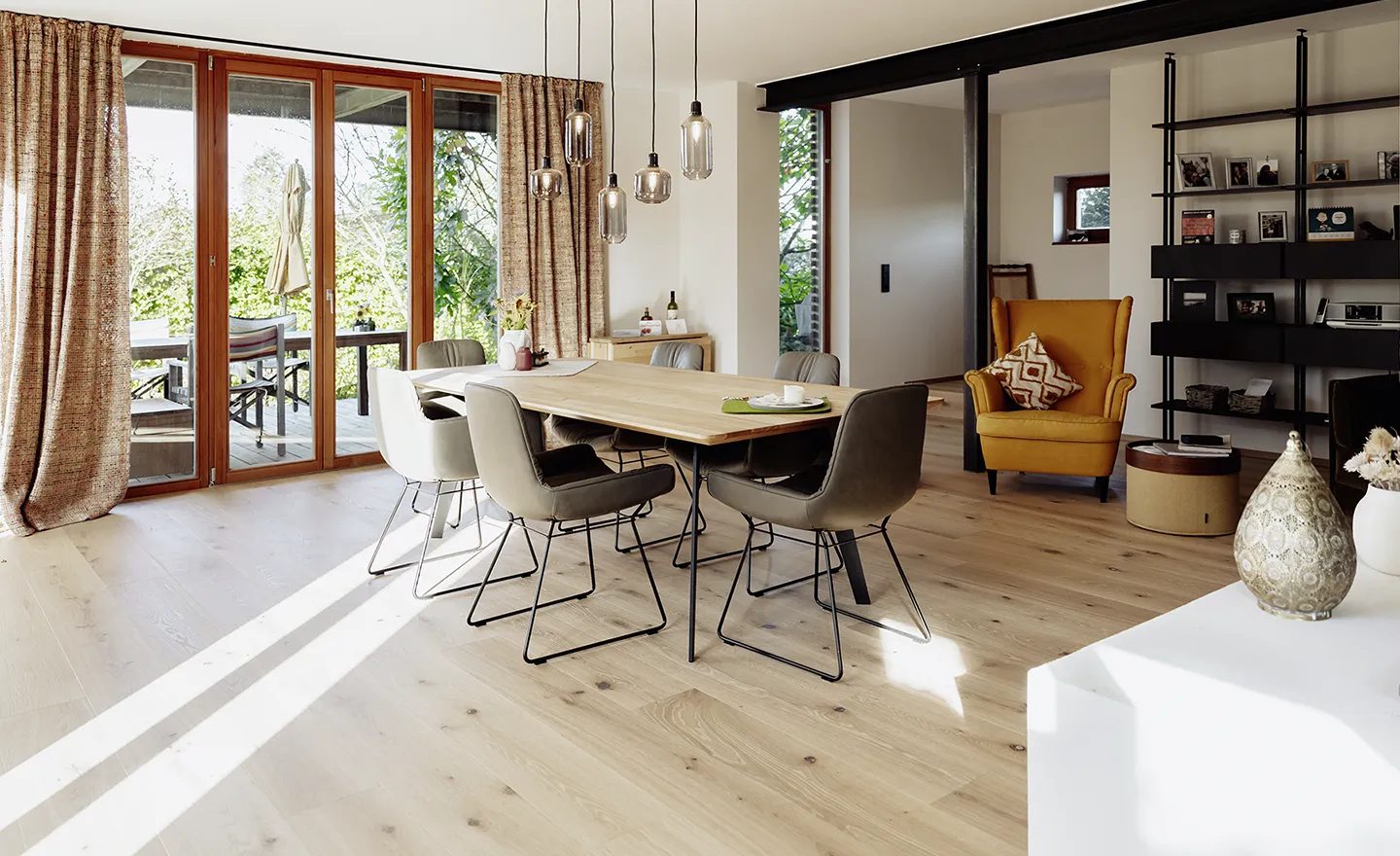 Heller Essbereich mit einem Holztisch, umgeben von sechs modernen Stühlen, Pendelleuchten, einem senffarbenen Sessel und großen Glastüren, die sich zu einer Außenterrasse öffnen.