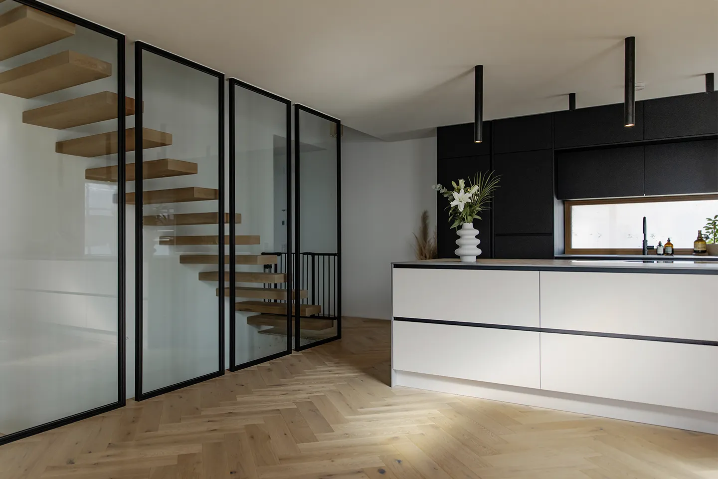 Moderne Küche mit weißer Kochinsel, schwarzen Schränken, freitragender Holztreppe hinter Glaspaneelen und einer Vase mit weißen Blumen auf der Kochinsel.