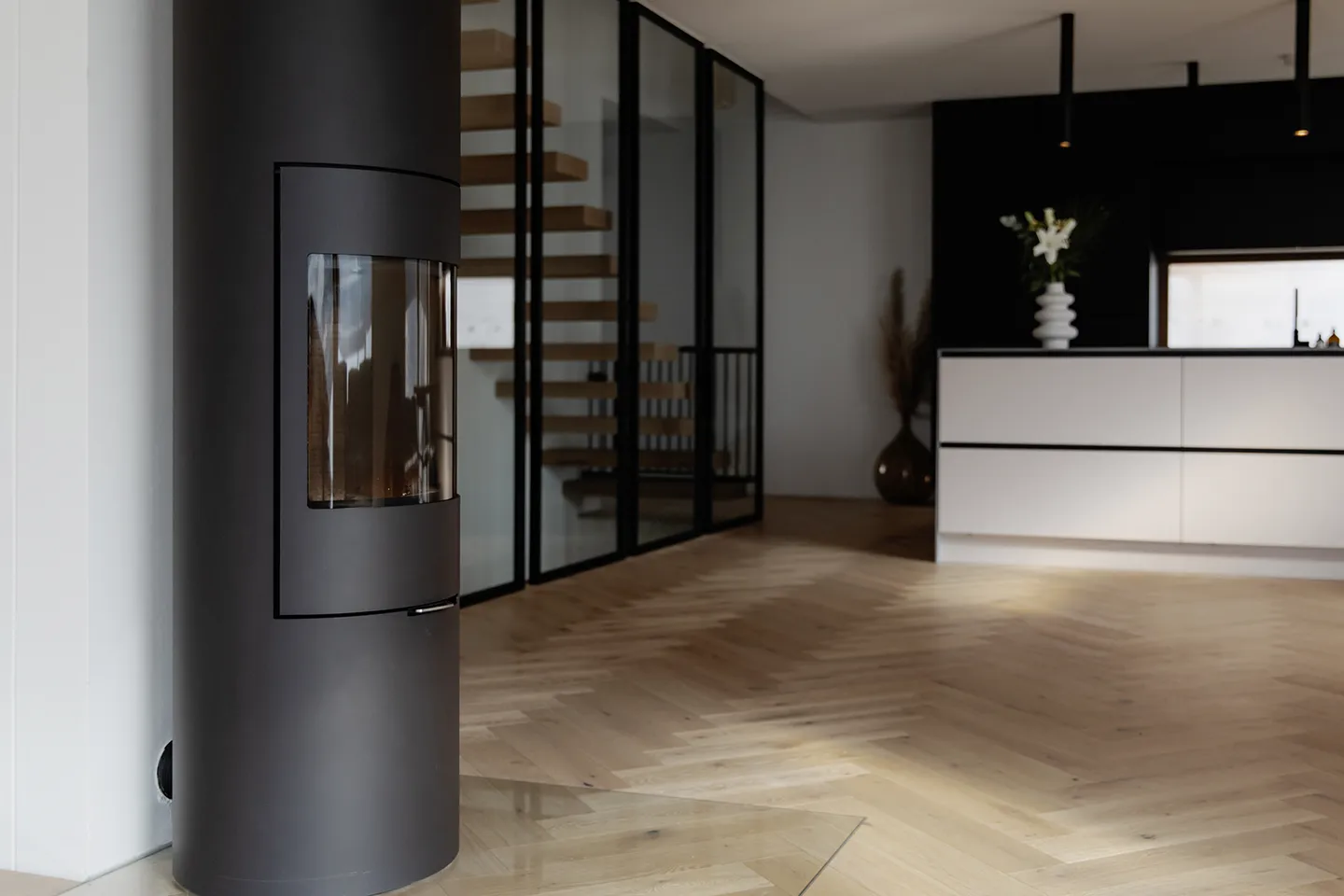 Moderner schwarzer zylindrischer Holzofen neben einer Glaswand mit Holztreppe in einem Raum mit hellem Fischgrätparkett und weißen Küchenschränken.