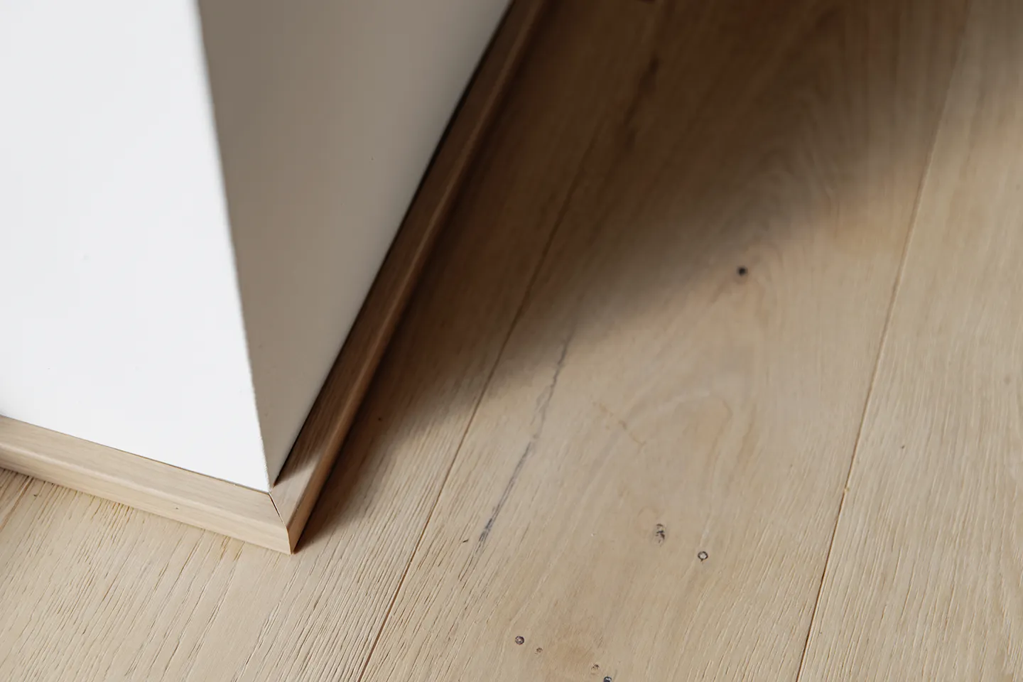 Ecke einer weißen Wand mit heller Holzfußleiste auf hellen Holzdielen mit natürlicher Maserung und Astlöchern.