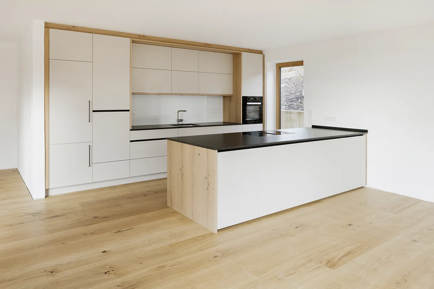 Moderne Küche mit hellem Holzboden, weißen Schränken, schwarzen Arbeitsplatten und einer Kochinsel mit eingebautem Herd.