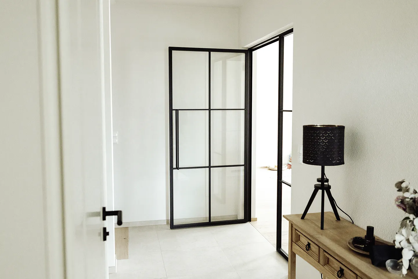 Minimalistischer Flur mit schwarz gerahmten Glastüren, hellem Fliesenboden, Holztisch und schwarzer Stativlampe.