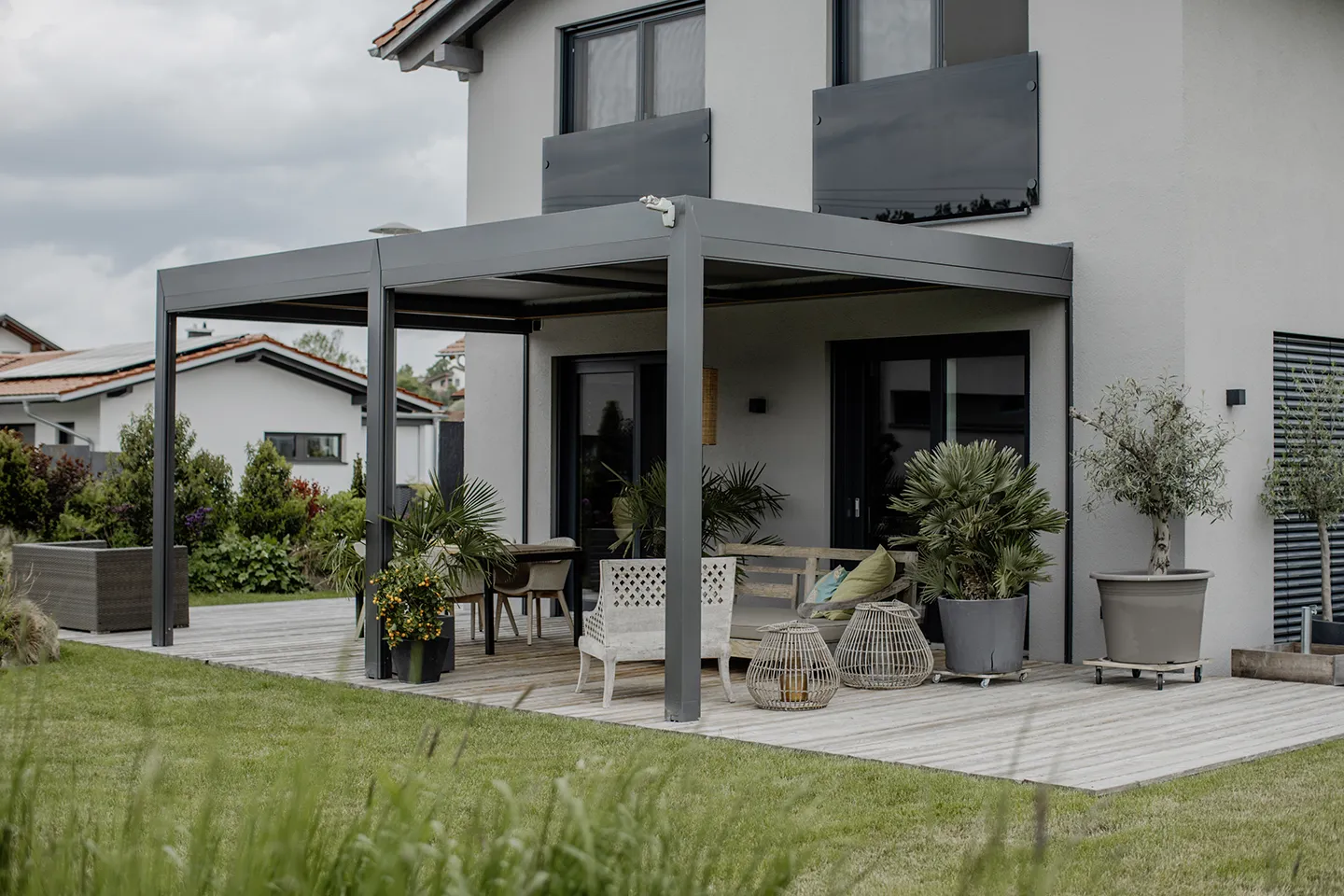 Moderne Terrasse mit grauer Pergola, Holzdeck, Sitzgelegenheiten im Freien, Topfpflanzen und einem grünen Rasen vor einem hellen Haus.