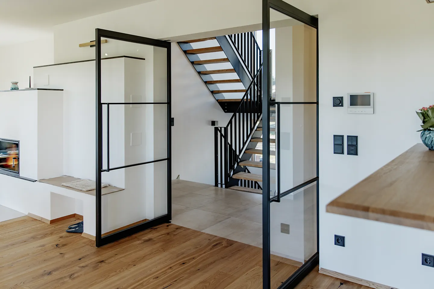 Modernes Interieur mit Holzboden, Glastüren mit schwarzen Rahmen, weißen Wänden und einer Treppe mit Holzstufen und schwarzem Geländer.