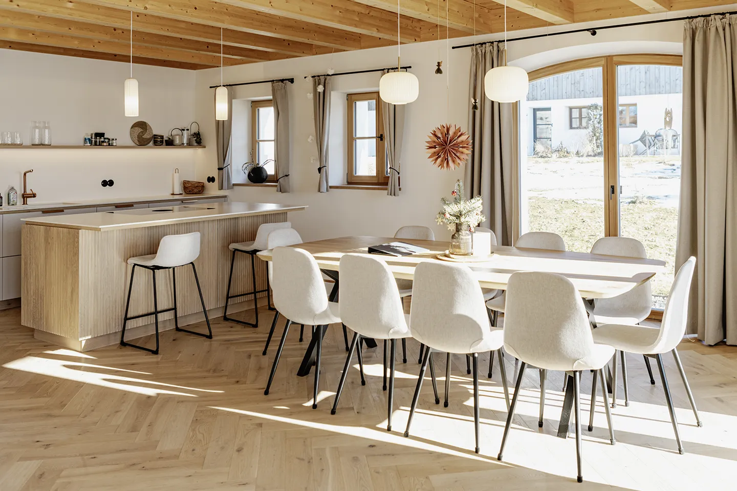 Helle, moderne Küche und Essbereich mit Holzdecke, großen Fenstern, hellen Holzböden, einem Esstisch mit acht weißen Stühlen und einer Kücheninsel mit zwei Barhockern.