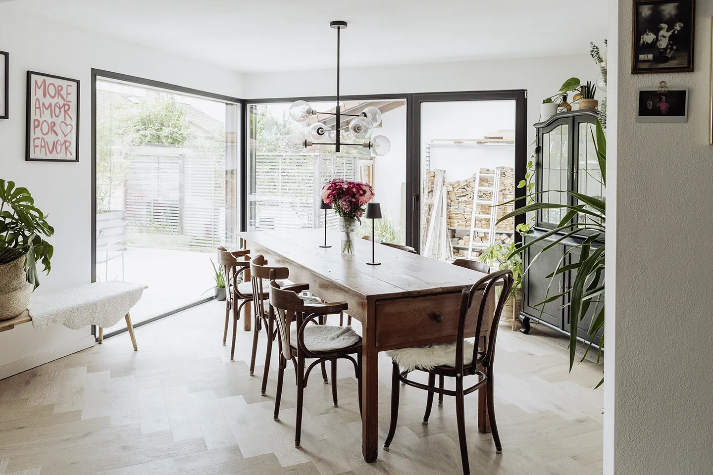 Helles Esszimmer mit einem großen Holztisch, umgeben von sechs Stühlen, einer Vase mit Blumen als Mittelpunkt, einem modernen Kronleuchter und großen Fenstern mit Blick in den Garten.