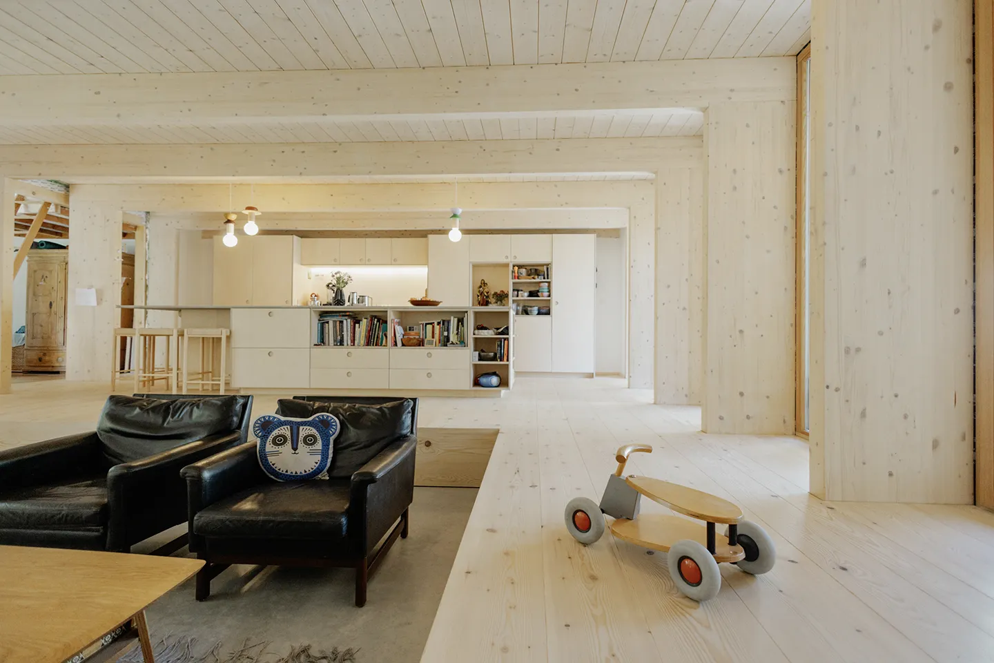 Moderner, offener Wohnbereich mit hellen Holzwänden, Decke und Boden, ausgestattet mit zwei schwarzen Ledersesseln, einem blau gemusterten Kissen und einem hölzernen Spielzeugauto auf Rädern.