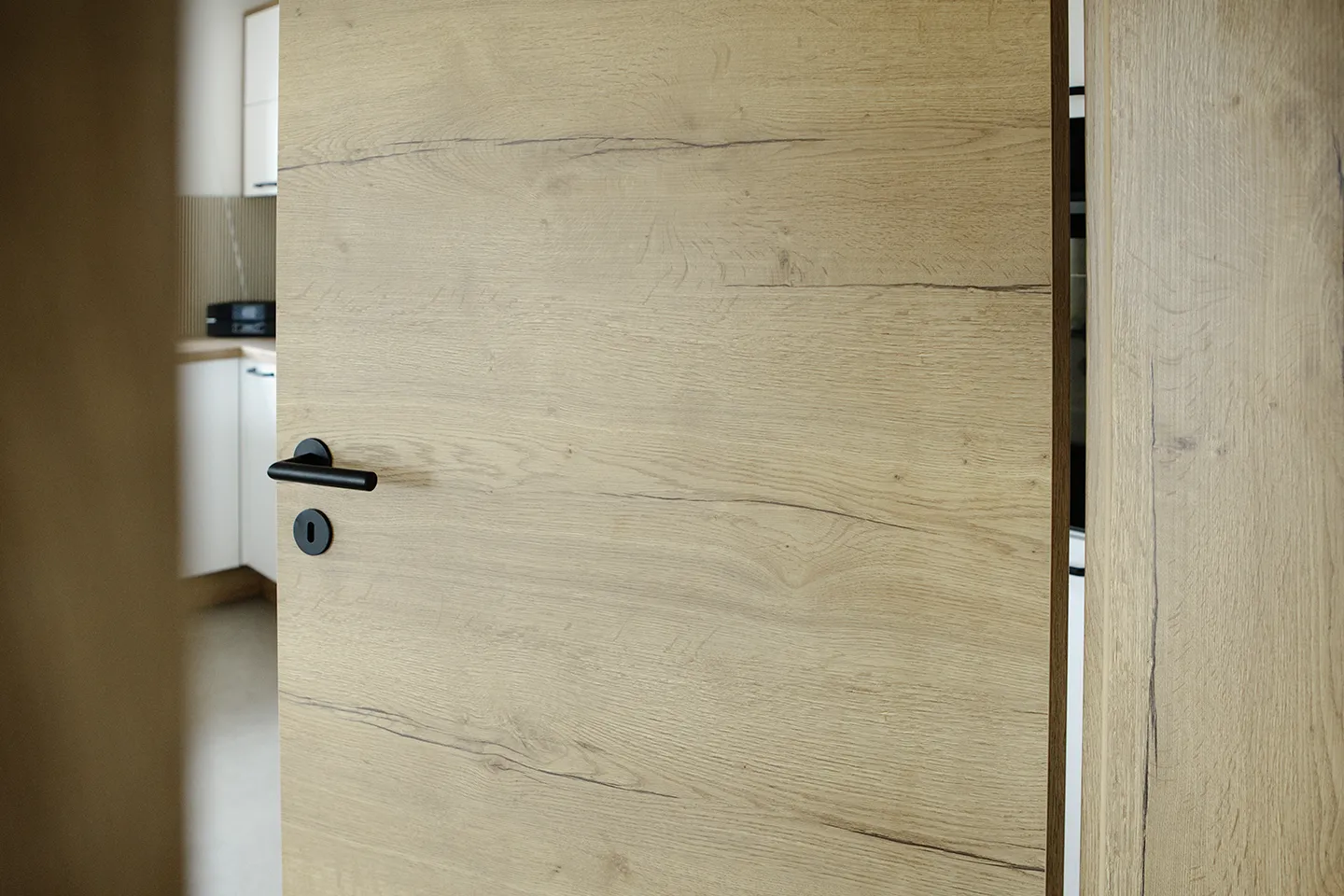Eine helle Holztür mit schwarzem Griff ist leicht geöffnet und gibt den Blick auf einen Teil des modernen Kücheninterieurs frei.