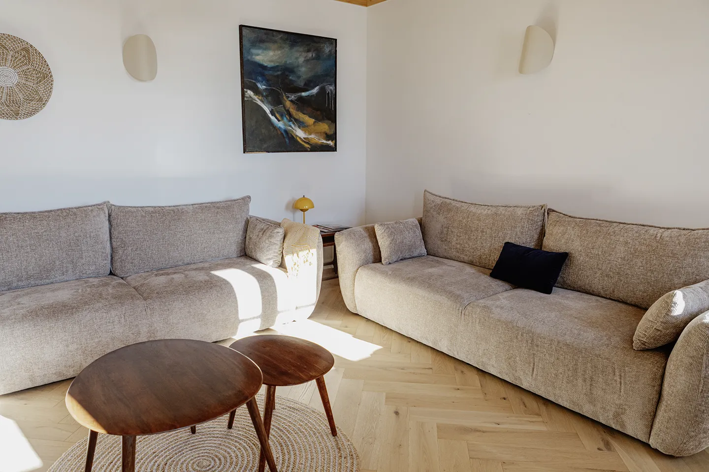 Eine Wohnzimmerecke mit zwei beigefarbenen Stoffsofas, einem runden Couchtisch aus Holz und darüber hängender abstrakter Wandkunst.