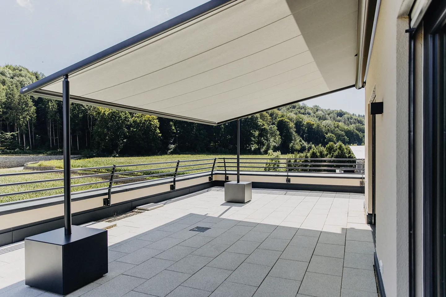 Großzügige, moderne Terrasse mit quadratischen Fliesen, Metallgeländer und einer großen, beigefarbenen, ausfahrbaren Markise mit Blick ins Grüne und auf Bäume.
