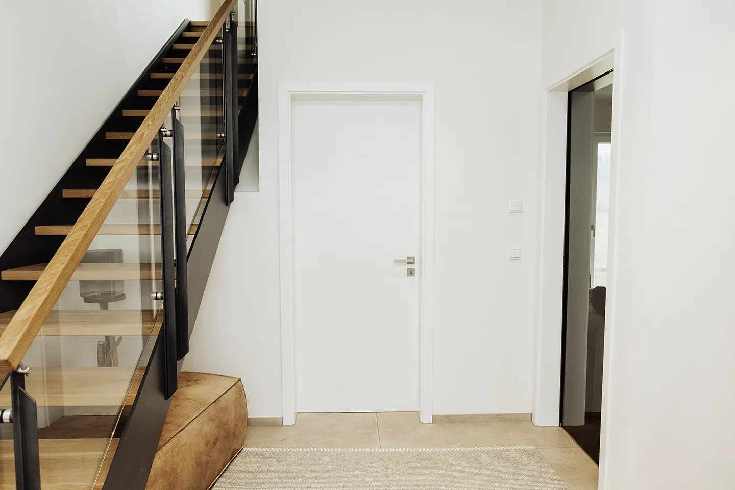 Minimalistischer Flur mit Holztreppe links, einer geschlossenen weißen Tür in der Mitte und einem offenen Durchgang rechts.