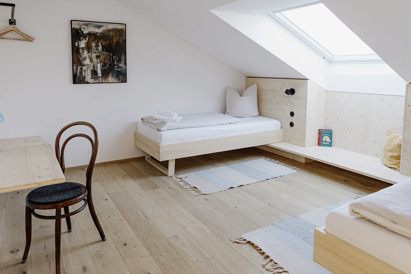 Minimalistisches Dachgeschoss-Schlafzimmer mit zwei Einzelbetten aus Holz, weißer Bettwäsche, Holzstuhl, Schreibtisch, Dachfenster und hellem Holzboden.