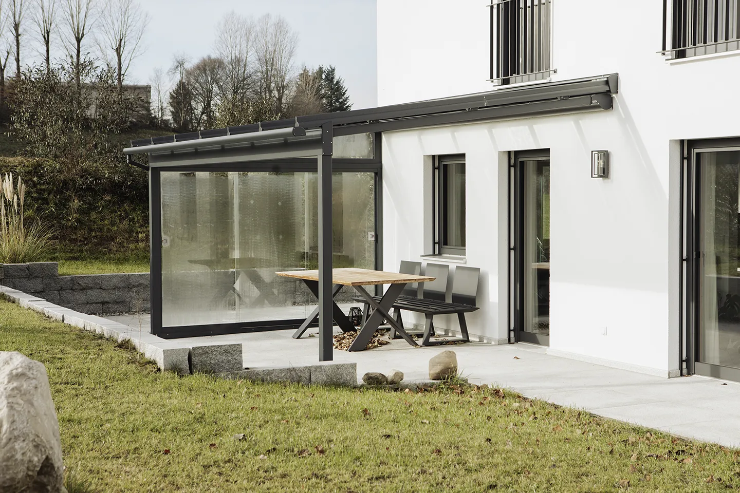 Moderne Hausterrasse mit verglastem Sitzbereich, Holztisch und Bank auf einer von Rasen umgebenen Betonplatte.