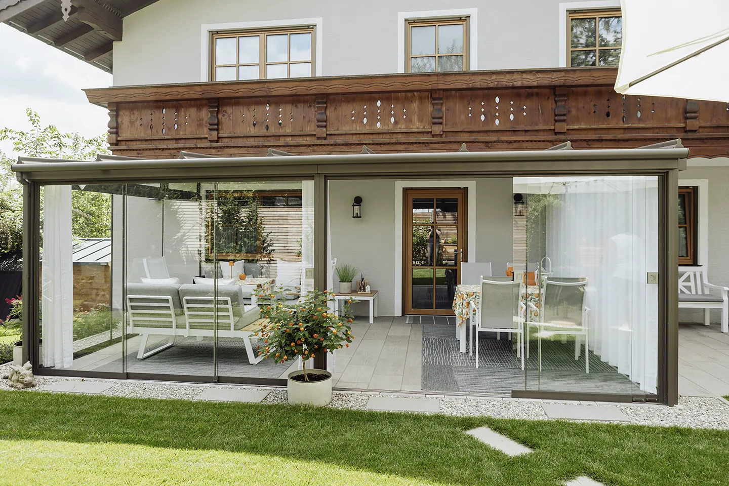 Moderner, verglaster Patio mit weißen Sitz- und Essbereichen im Freien vor einem Haus mit Holzbalkon.