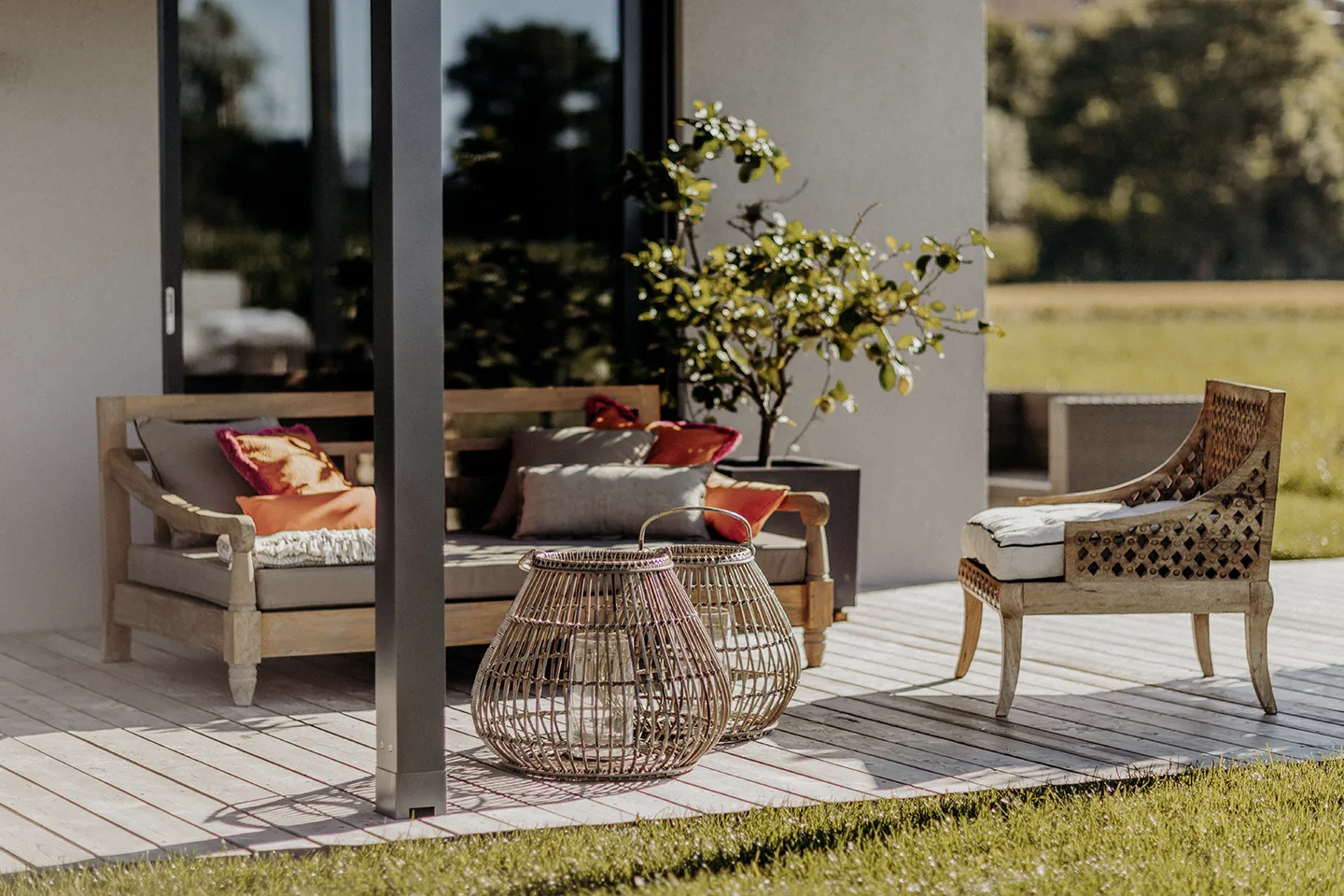 Terrasse im Freien mit gepolstertem Holzsofa und -stuhl, Korblaternen und einer Topfpflanze auf einem Holzdeck.