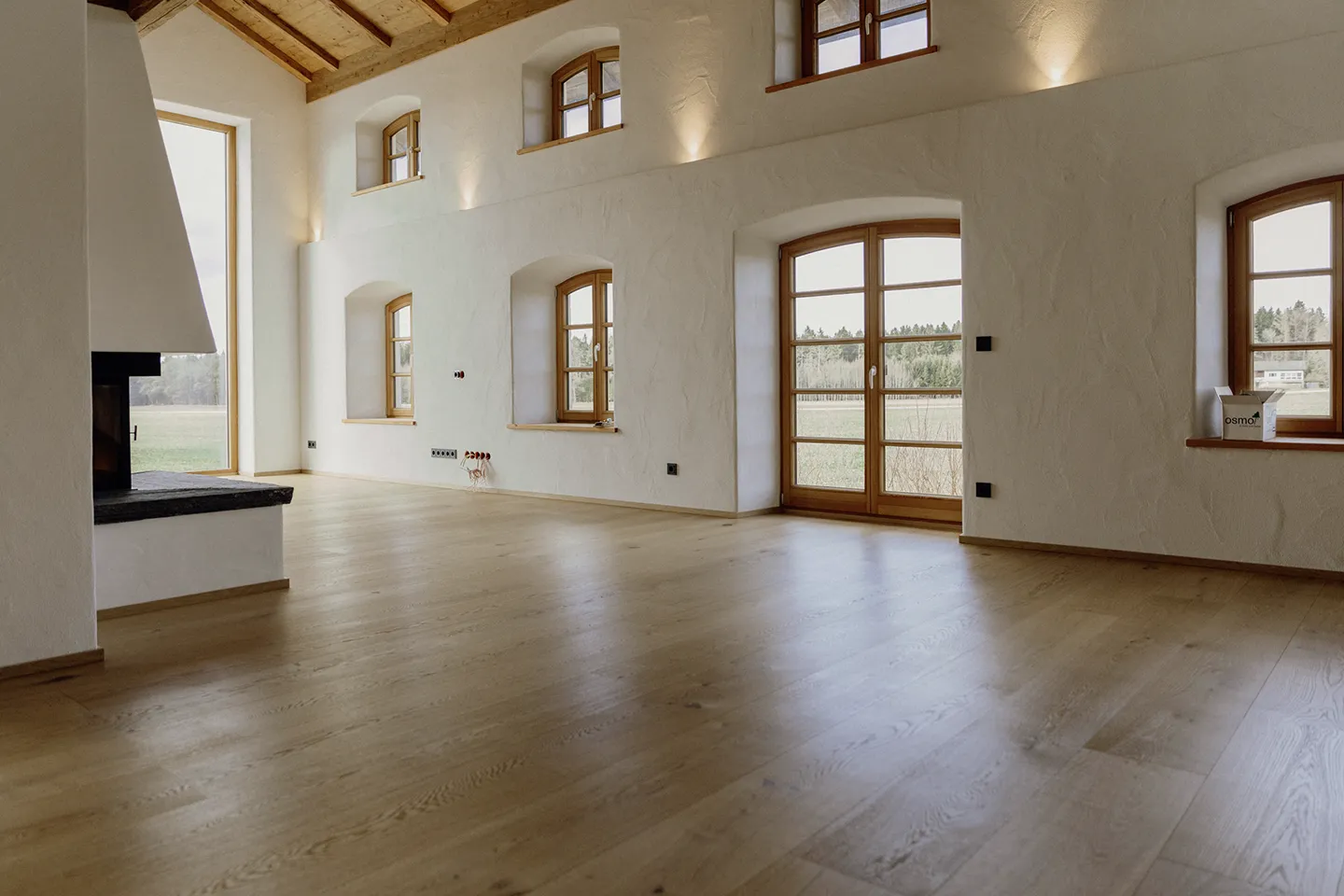 Großzügiger, leerer Raum mit Holzboden, weiß strukturierten Wänden, Holzbalken an der Decke, mehreren Bogenfenstern und einem Kamin.