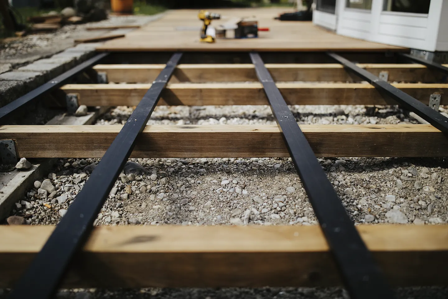 Holzrahmenkonstruktion auf Kiesgrund und im Hintergrund eine Bohrmaschine in der Nähe eines weißen Gebäudes.