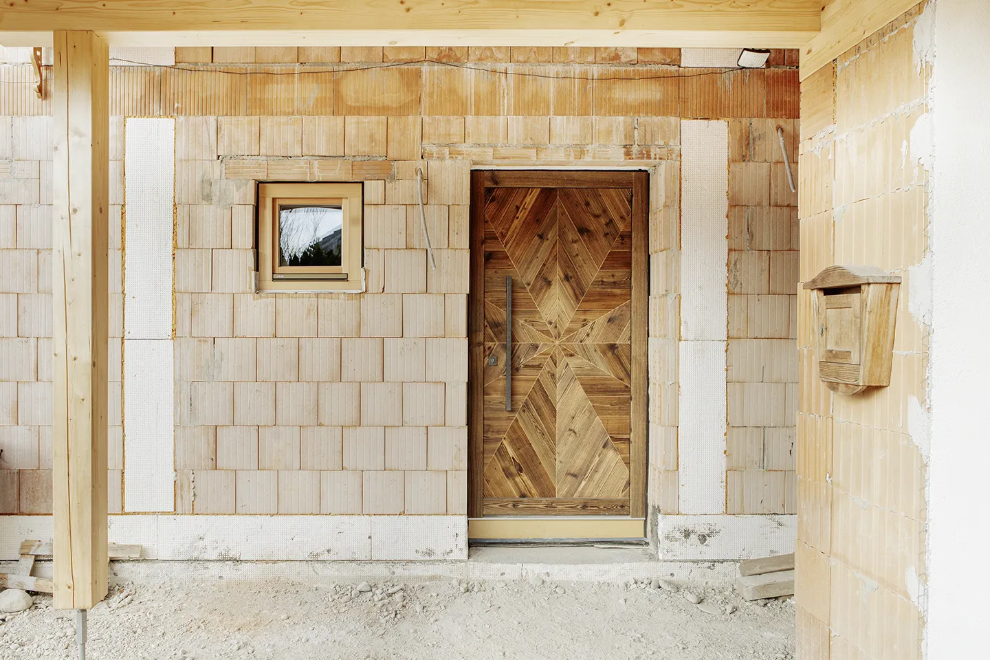 Unfertiger Backsteinhauseingang mit dekorativer Holzeingangstür, kleinem Fenster und hölzernem Briefkasten.