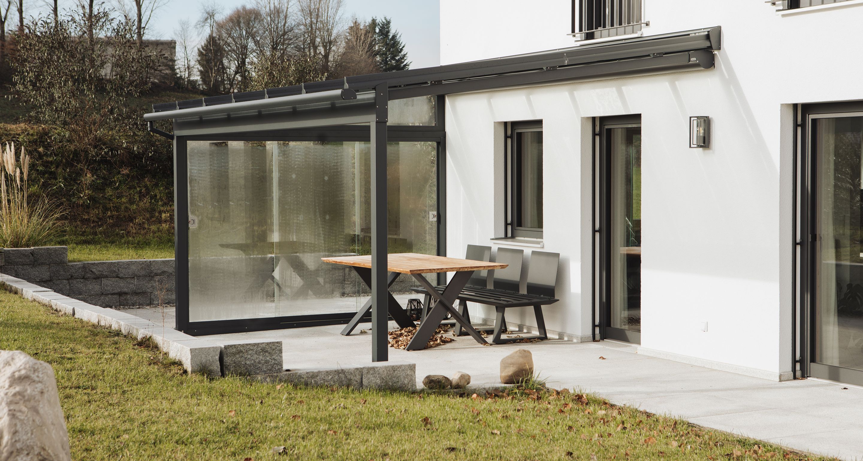 Modernes Terrassendach aus Aluminium mit Glaseindeckung und seitlichen Glasschiebeelements als Windschutz. Darunter ein Gartentisch mit Stühlen auf einer gefliesten Terrasse.