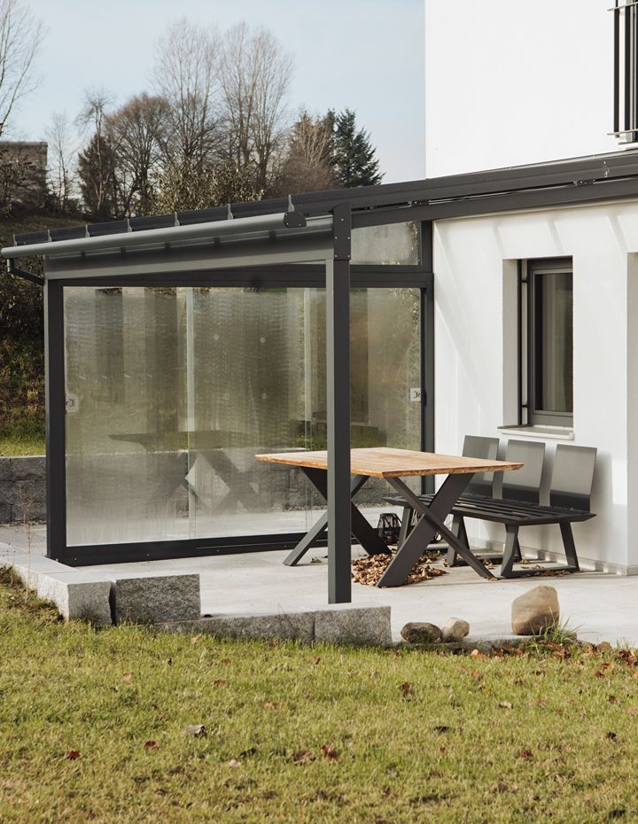 Modernes Terrassendach aus Aluminium mit Glaseindeckung und seitlichen Glasschiebeelements als Windschutz. Darunter ein Gartentisch mit Stühlen auf einer gefliesten Terrasse.