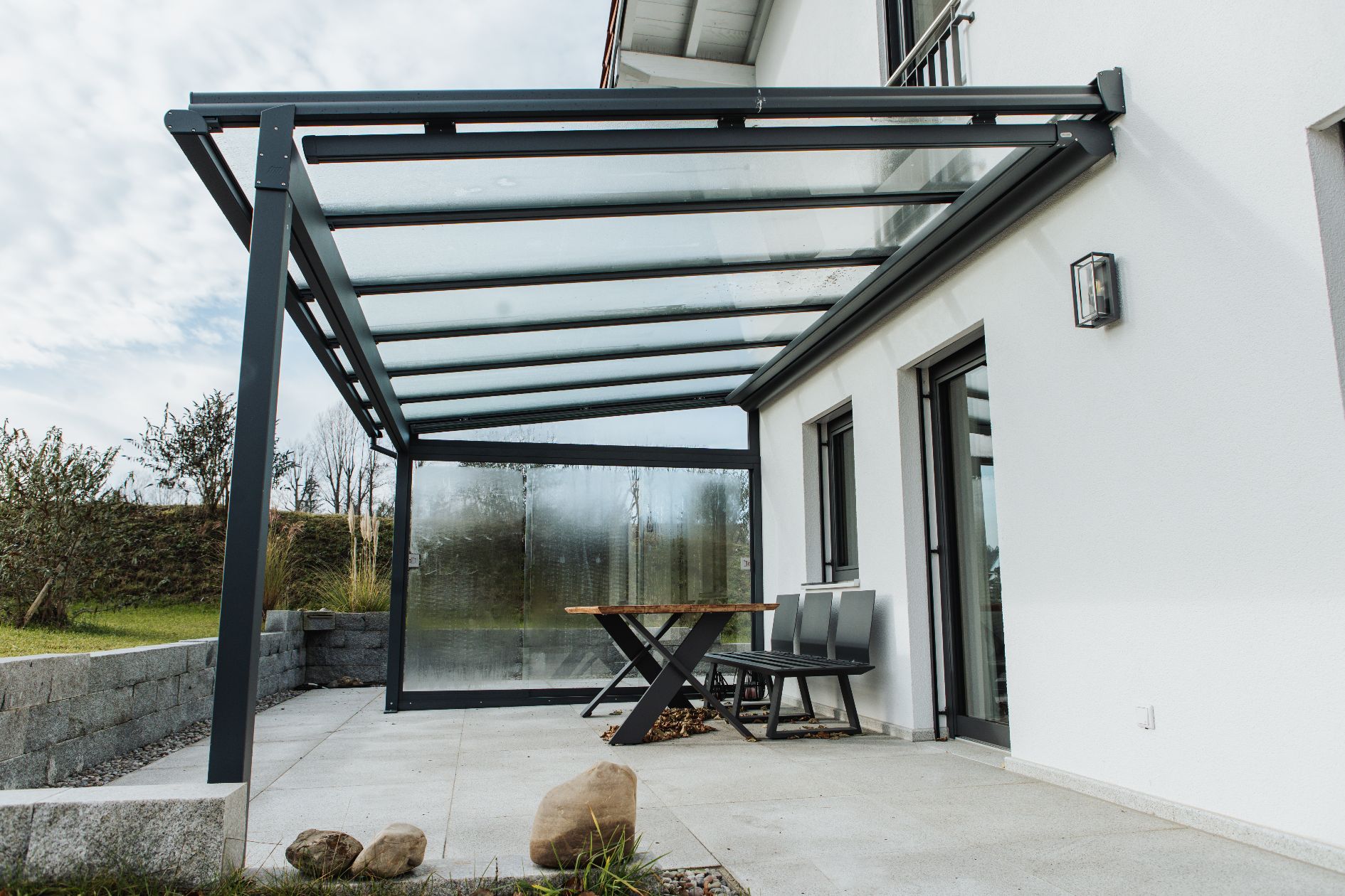 Terrassendach mit Glaseindeckung, anthrazitfarbenem Aluminiumgestell und seitlichem Windschutz aus Schiebeelementen.