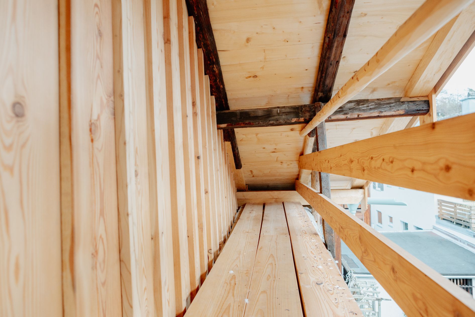 Detailaufnahme des neu aufgebauten Holzbalkons mit Lärchenboden und Geländer, im Kontrast zu den originalen, dunklen Dachbalken.