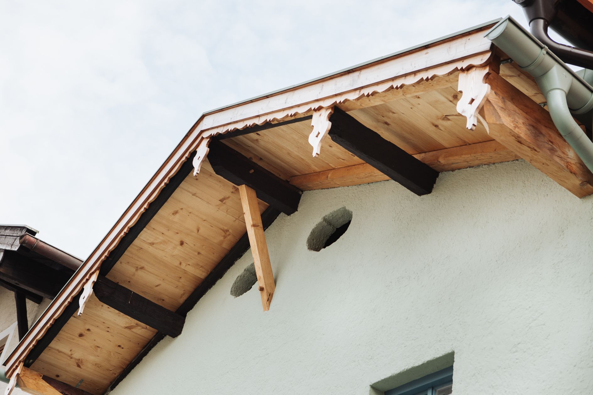 Holzdachüberstand mit geschnitzten Zierbrettern und Balken an verputzter Hausfassade.
