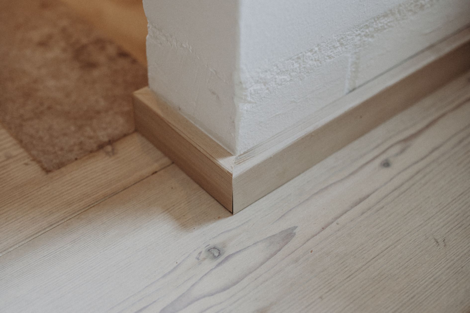 Detailaufnahme von Sockelleiste auf weiß geölter Lärchendiele an weiß verputzter Wand.