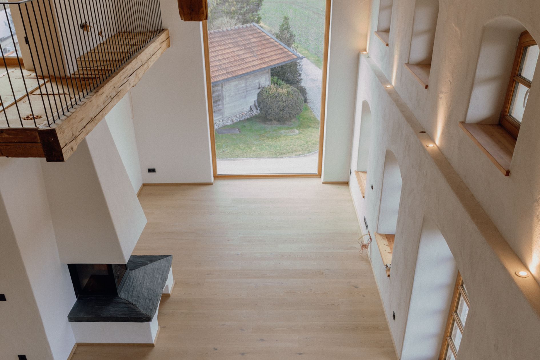 Blick von oben auf einen großzügigen Wohnraum mit geöltem Eichenboden, Kamin und raumhohem Fenster mit Blick ins Grüne.