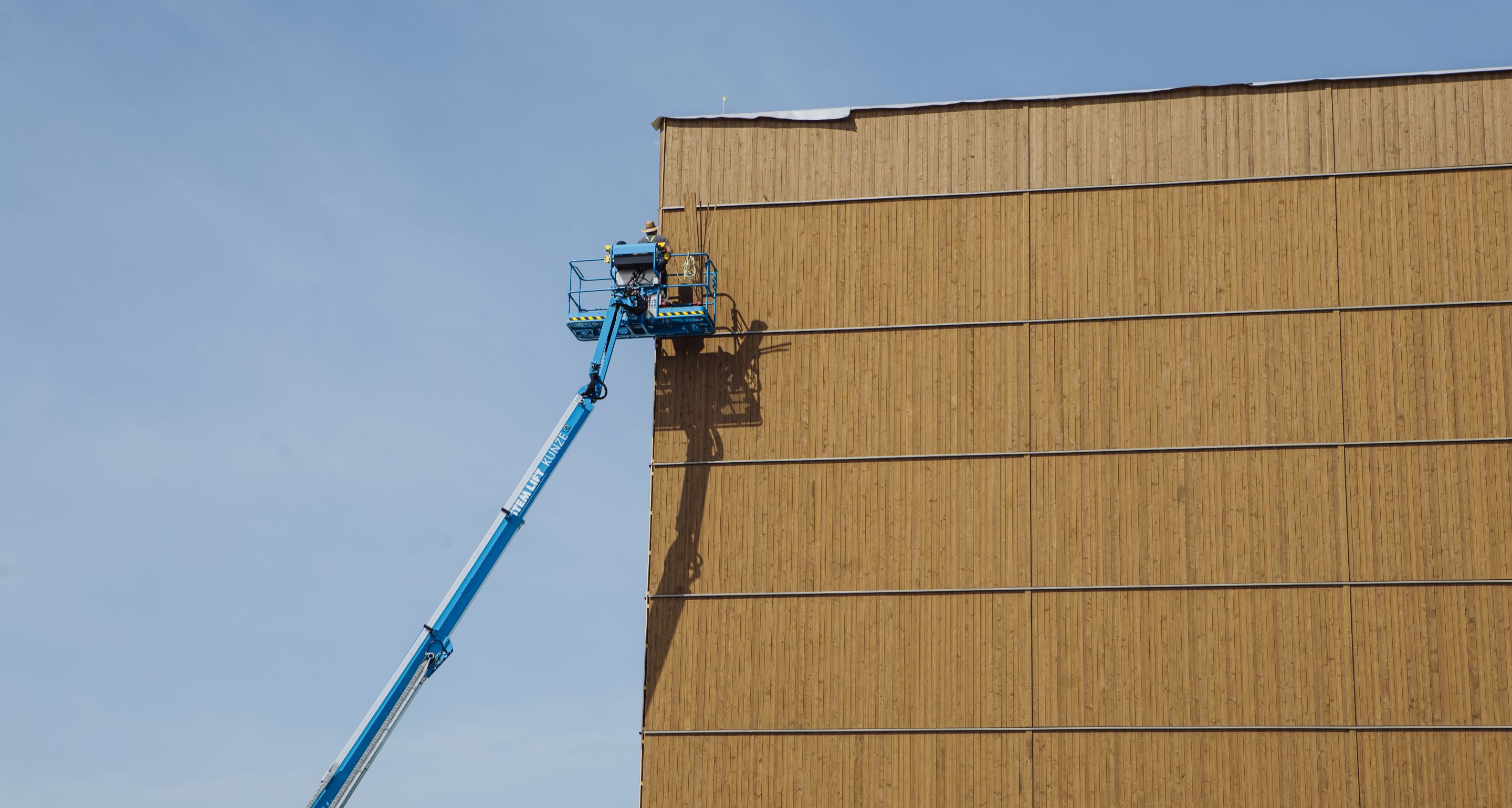 Arbeiter auf Hebebühne montieren vertikale Holzverkleidung an einer hohen Fassade